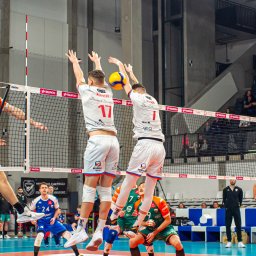 Tryb Manualny by Jakub Szołdra-Laszczyk - Mecz siatkówki: dwóch zawodników w białych strojach z numerami 17 i 1 blokuje atak przeciwnika, dynamiczna akcja nad siatką, publiczność na trybunach w tle.