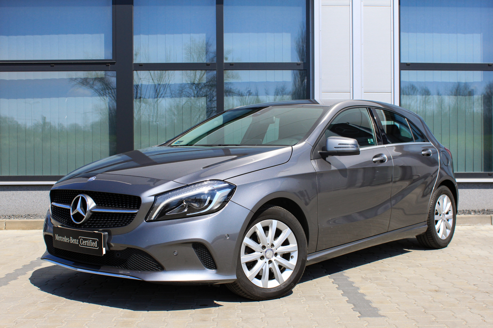Szary Mercedes-Benz Klasy A zaparkowany na zewnątrz budynku z dużymi oknami, z widocznym certyfikatem Mercedes-Benz.