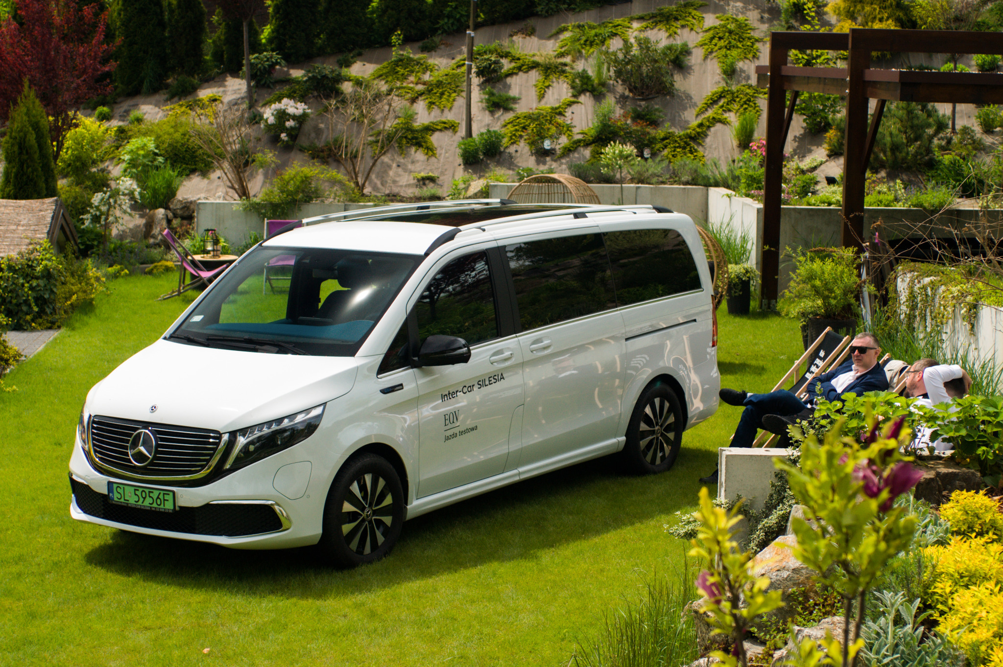 Biały Mercedes-Benz EQV z napisem Inter-Car Silesia na tle zielonego ogrodu z wypoczywającymi osobami w tle.