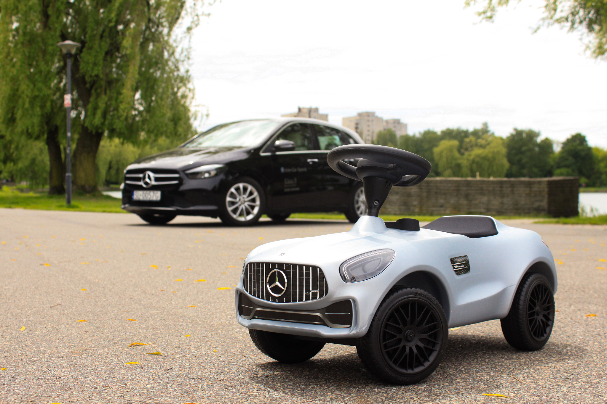 Jasnoniebieski jeździk dla dziecka stylizowany na Mercedesa AMG na tle zaparkowanego czarnego Mercedesa B-klasy w parku.