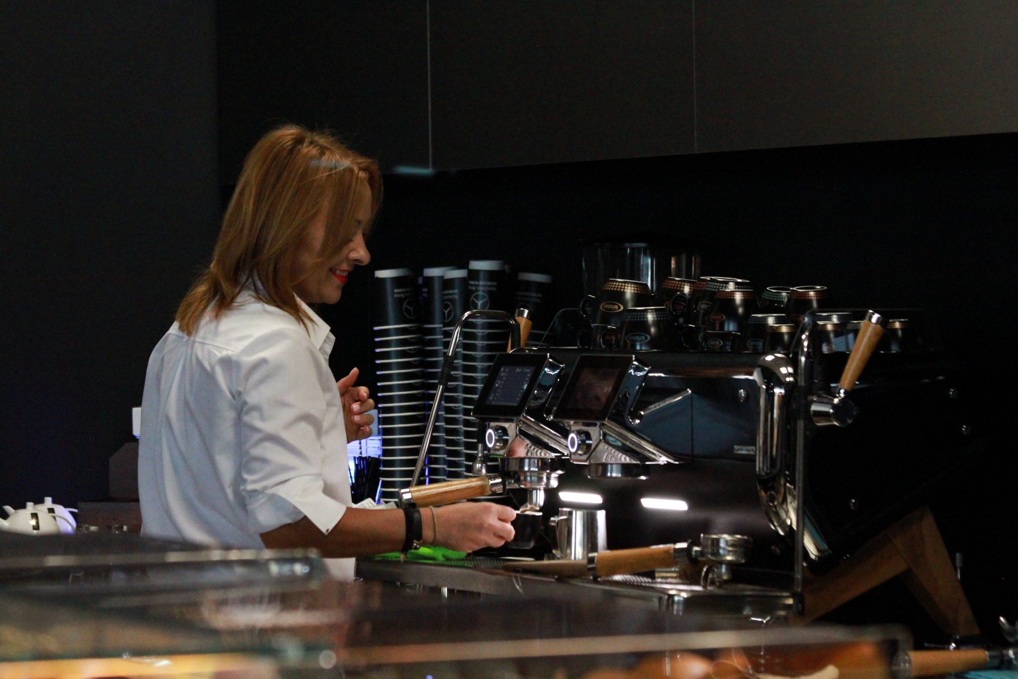 Kobieta w białej koszuli obsługuje profesjonalny ekspres do kawy, przygotowując napój. W tle widoczne kubki i filiżanki.
