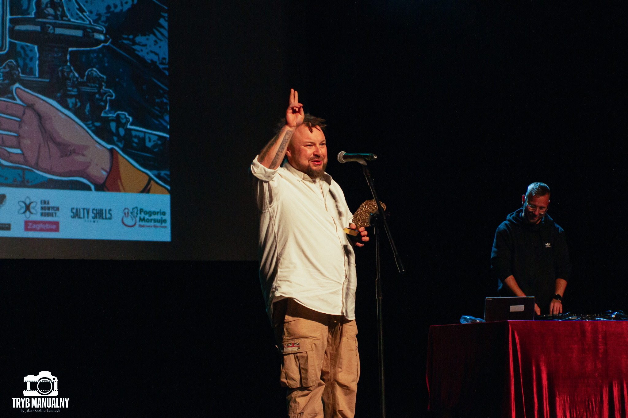 Mężczyzna na scenie z mikrofonem i nagrodą, w tle ekran z grafiką i drugi mężczyzna przy stole DJ-skim.