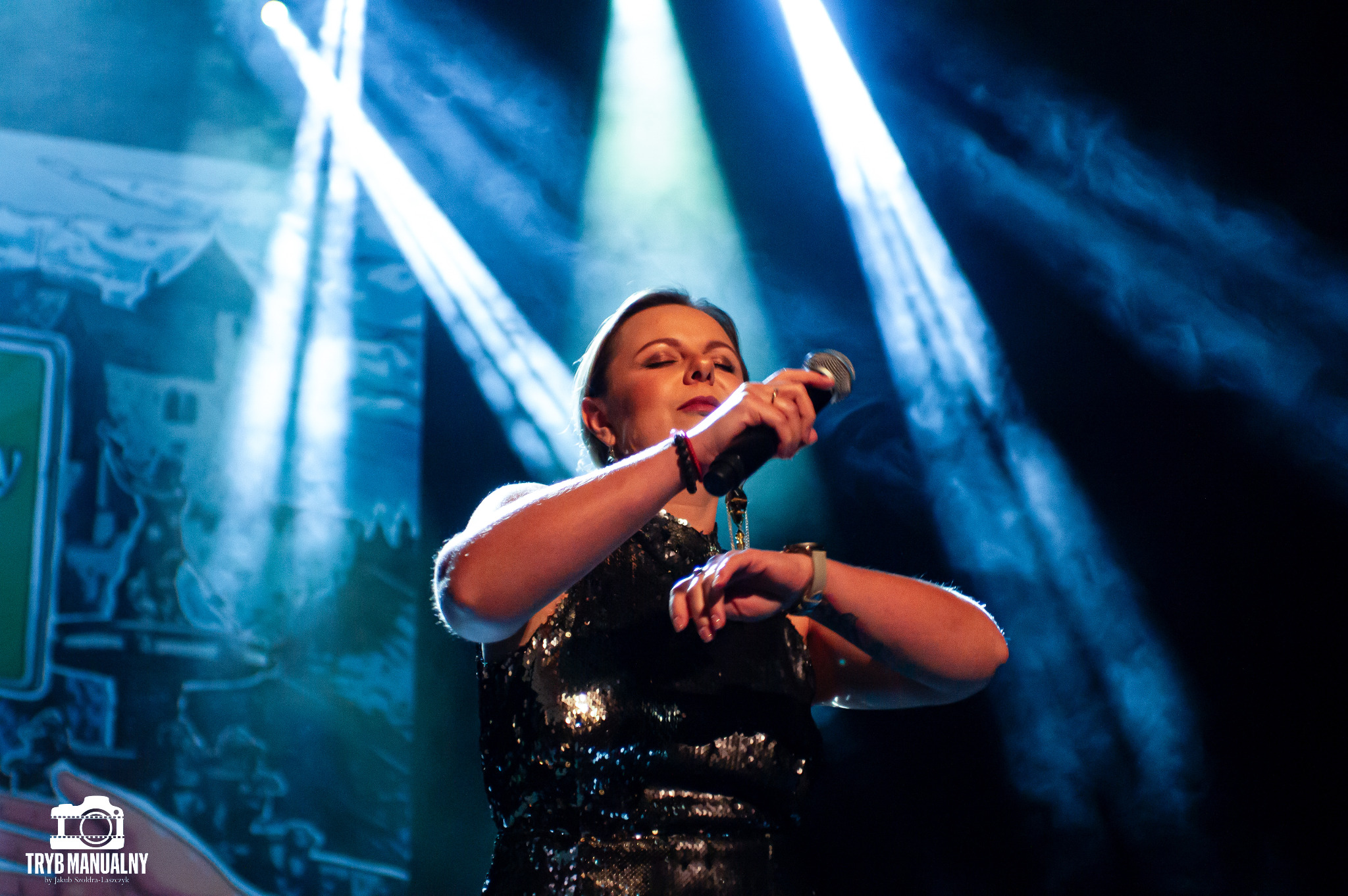 Wokalistka w błyszczącej sukience śpiewa na scenie, oświetlona reflektorami, z mikrofonem w dłoni i zamkniętymi oczami.