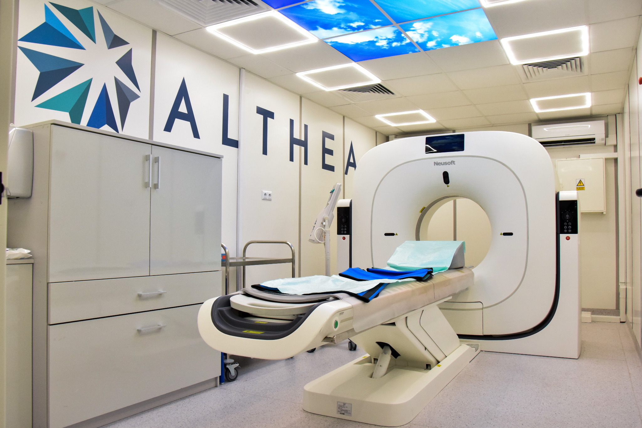 Nowoczesny skaner CT w jasnym, sterylnym pomieszczeniu z podświetlanym sufitem imitującym niebo, obok szafka i logo firmy Althea na ścianie.