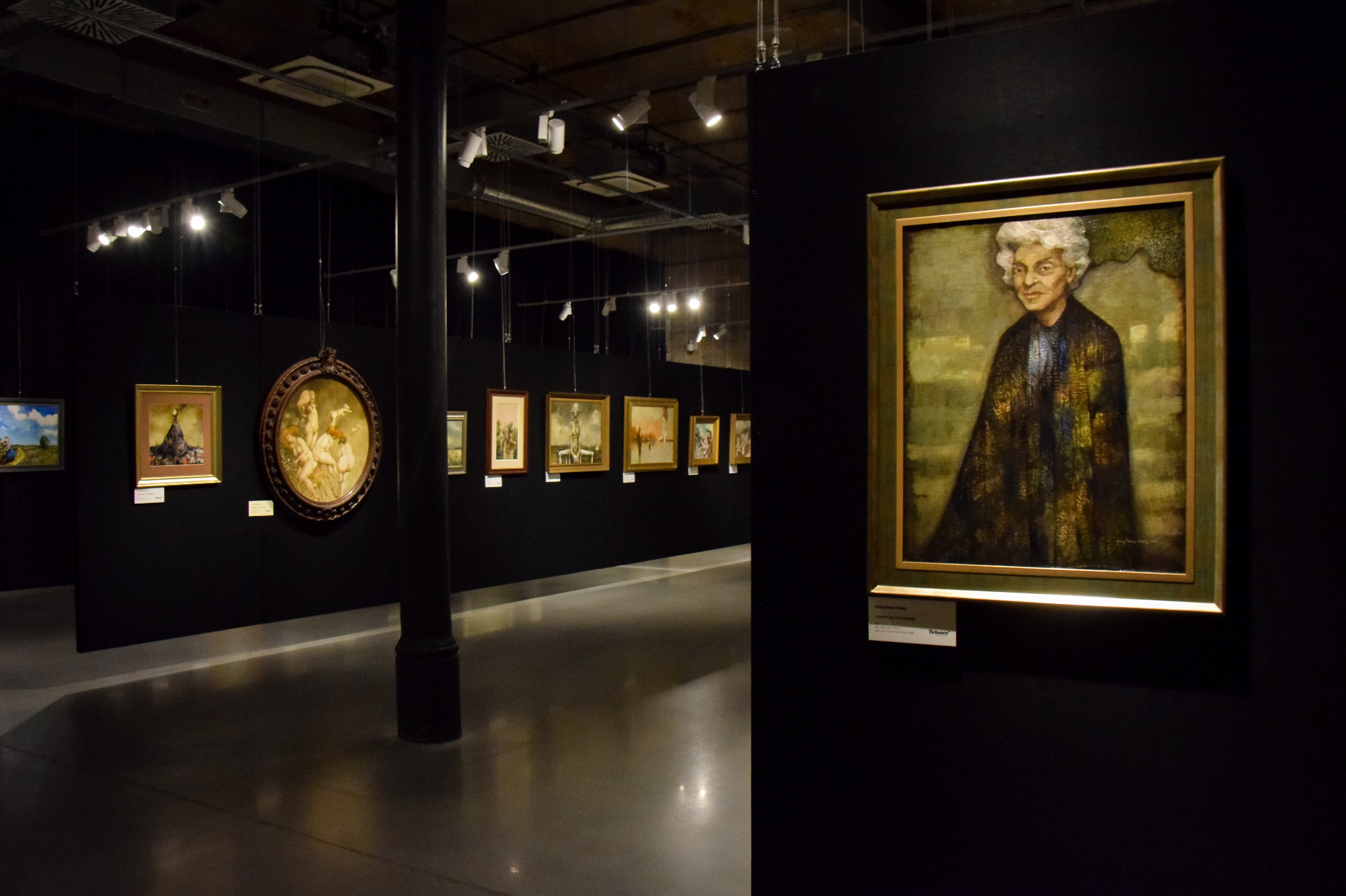 Wnętrze galerii sztuki z obrazami na czarnych ścianach, oświetlonych reflektorami, z polerowaną podłogą odbijającą światło. Na pierwszym planie portret starszej kobiety w złotej ramie.