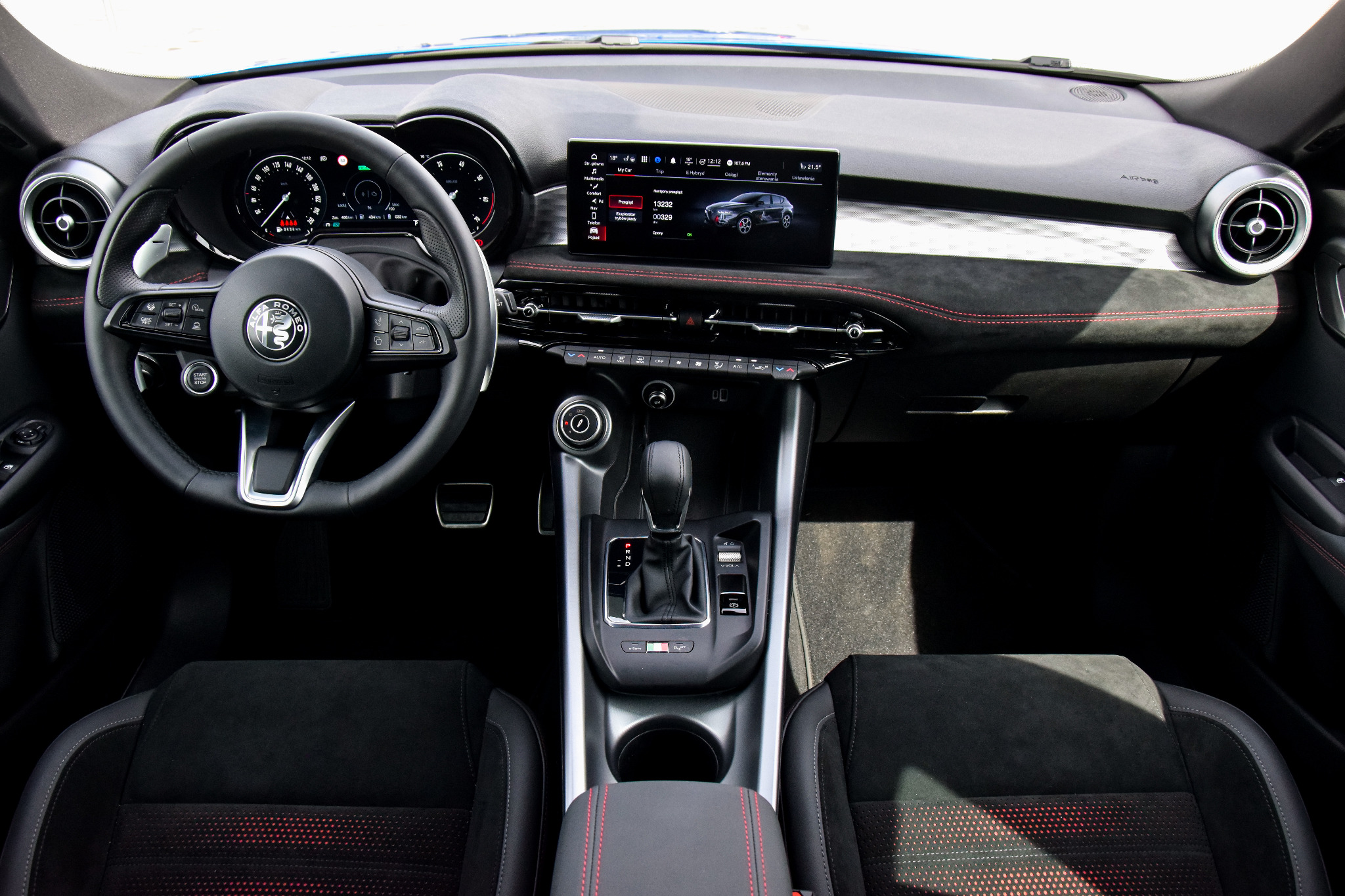 Wnętrze samochodu Alfa Romeo z czarną deską rozdzielczą obszytą czerwoną nicią, widoczny ekran nawigacji prezentujący model samochodu, kierownica z logo marki oraz automatyczna skrzynia biegów.