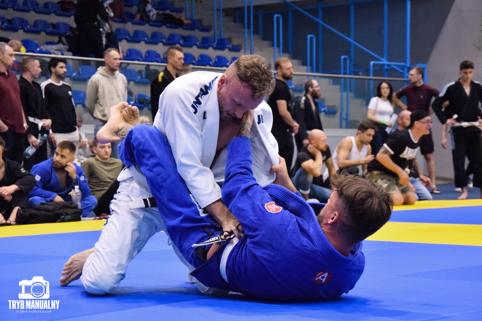 Dwóch mężczyzn w kimonach, białe i niebieskie, walczących na niebiesko-żółtej macie podczas zawodów brazylijskiego jiu-jitsu, z publicznością w tle.