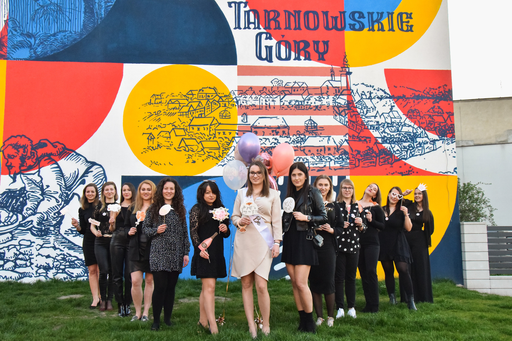 Grupa kobiet pozuje na tle kolorowego muralu z napisem Tarnowskie Góry, trzymając rekwizyty do zdjęć i balony, prawdopodobnie podczas wieczoru panieńskiego.