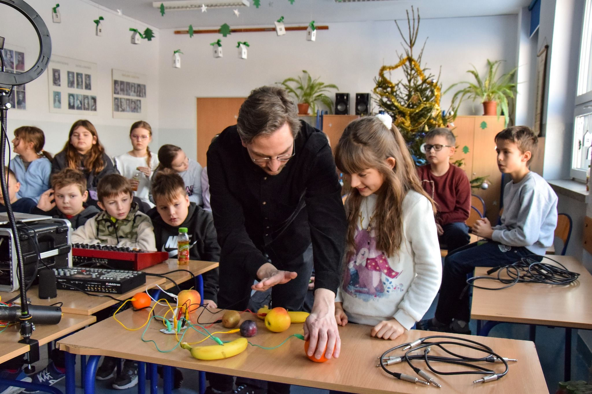 Nauczyciel demonstruje eksperyment z owocami i przewodami uczniom w klasie, na tle choinka i sprzęt audio.