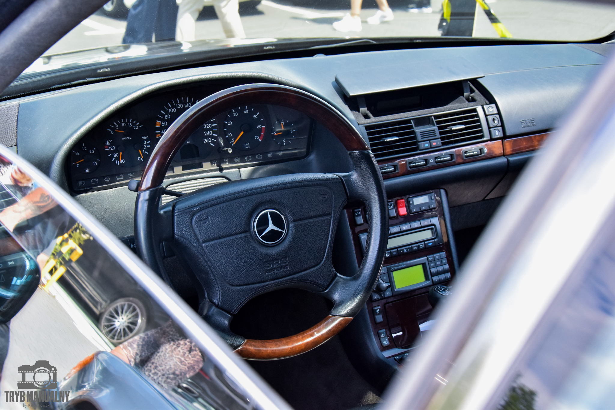 Wnętrze klasycznego samochodu Mercedes-Benz z drewnianą kierownicą, widok zza szyby, deska rozdzielcza z analogowymi zegarami i zielonym wyświetlaczem radia, detale wykończenia w drewnie.