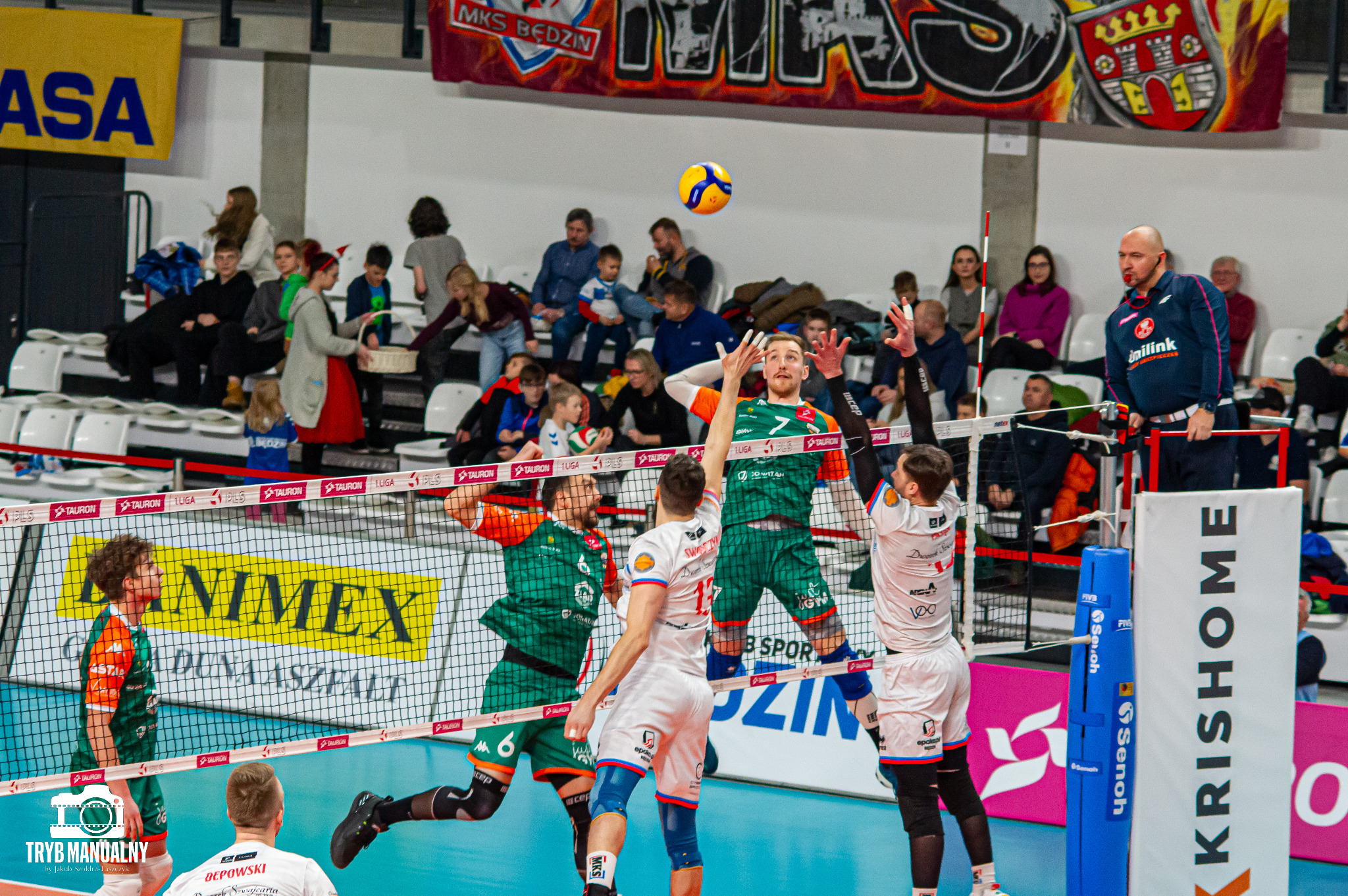 Akcja podczas meczu siatkówki halowej: zawodnicy w zielono-pomarańczowych i biało-niebieskich strojach walczą o piłkę nad siatką na tle trybun wypełnionych kibicami.