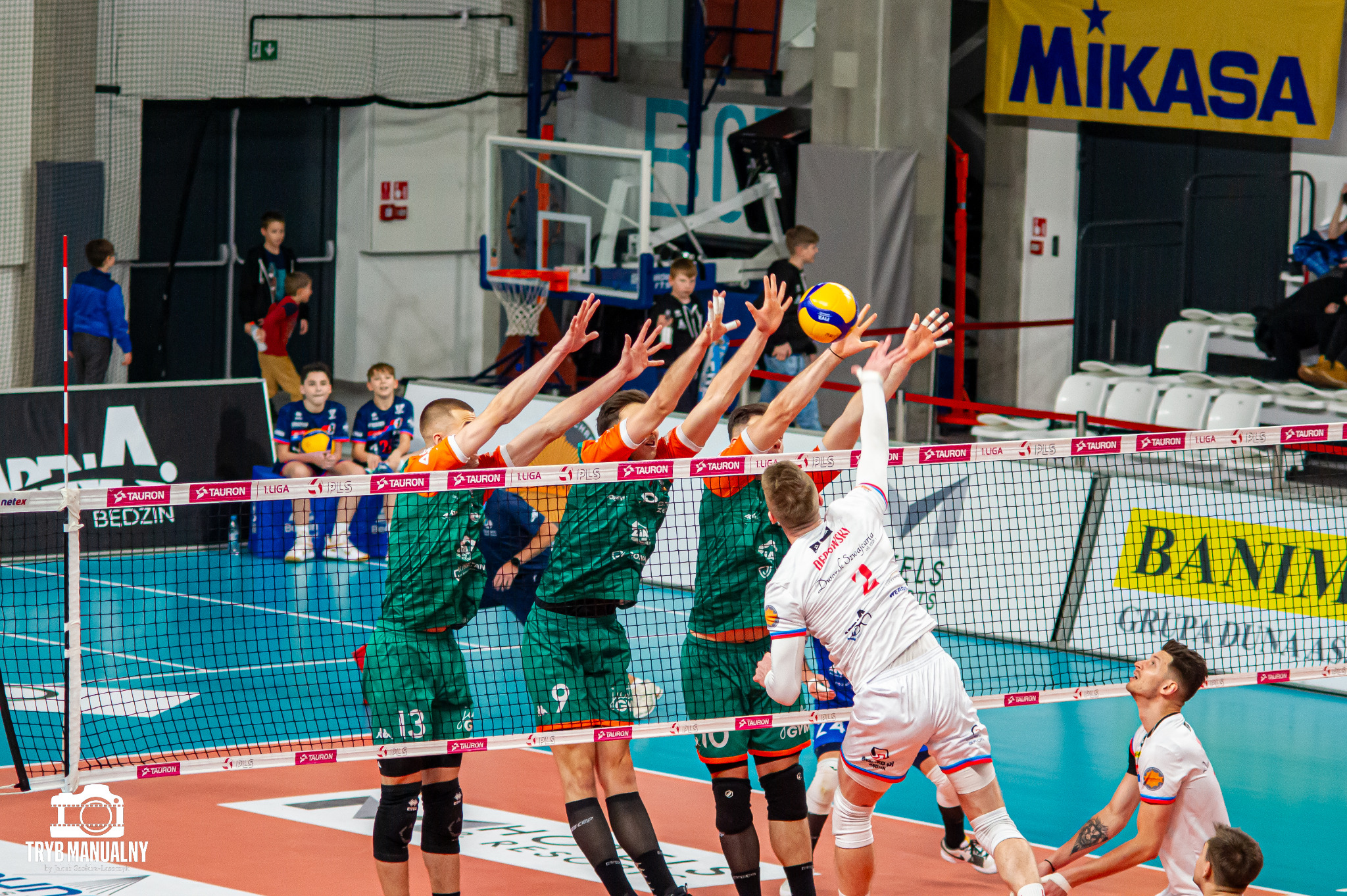 Mecz siatkówki: blok czterech zawodników w zielono-pomarańczowych strojach próbujących zatrzymać atakującego w białym stroju z numerem 2, w tle widownia i banery reklamowe.