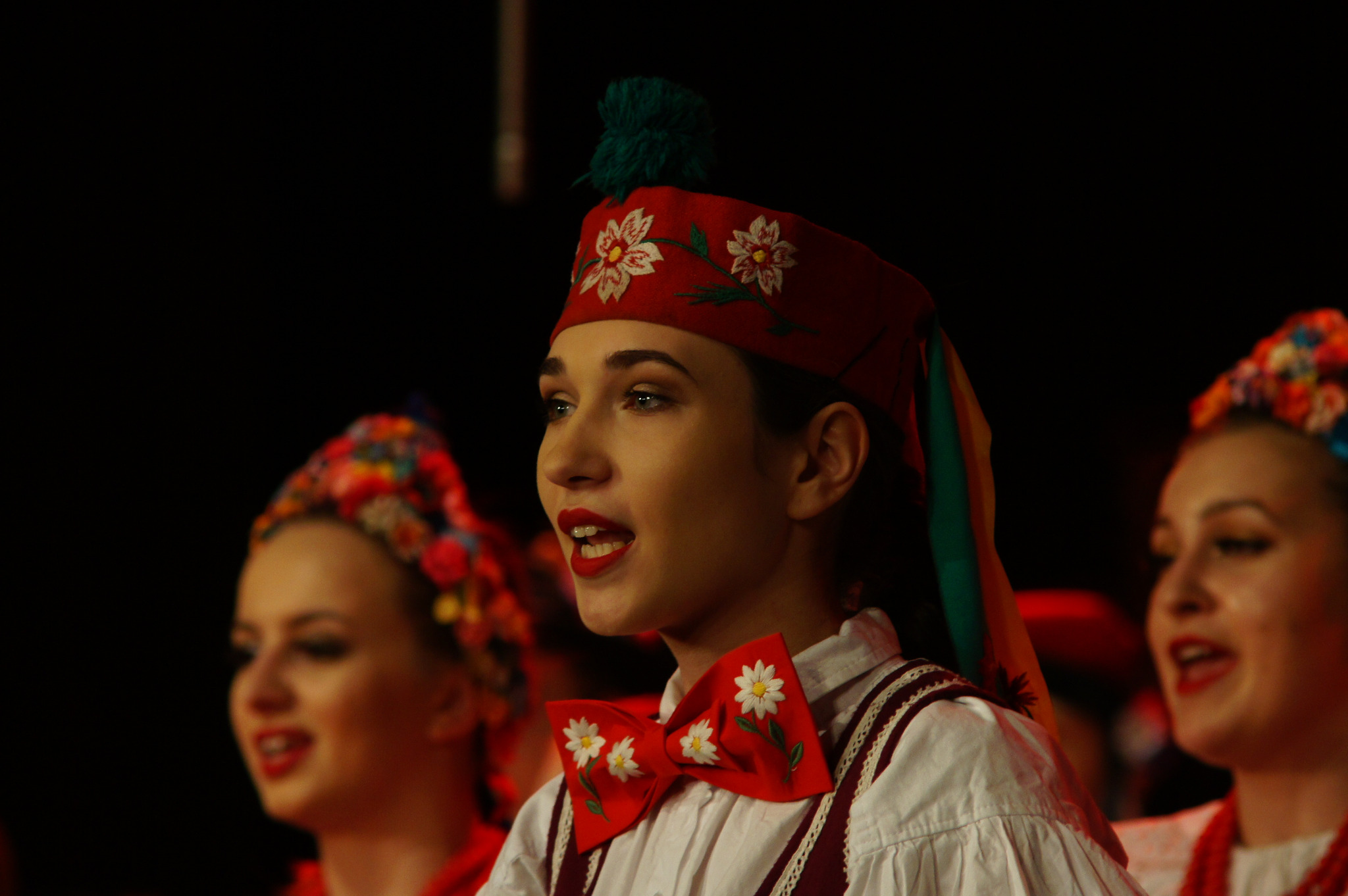 Młoda kobieta w tradycyjnym stroju ludowym, z czerwoną czapką z kwiatami i kokardą, śpiewa na scenie z innymi tancerkami w tle.