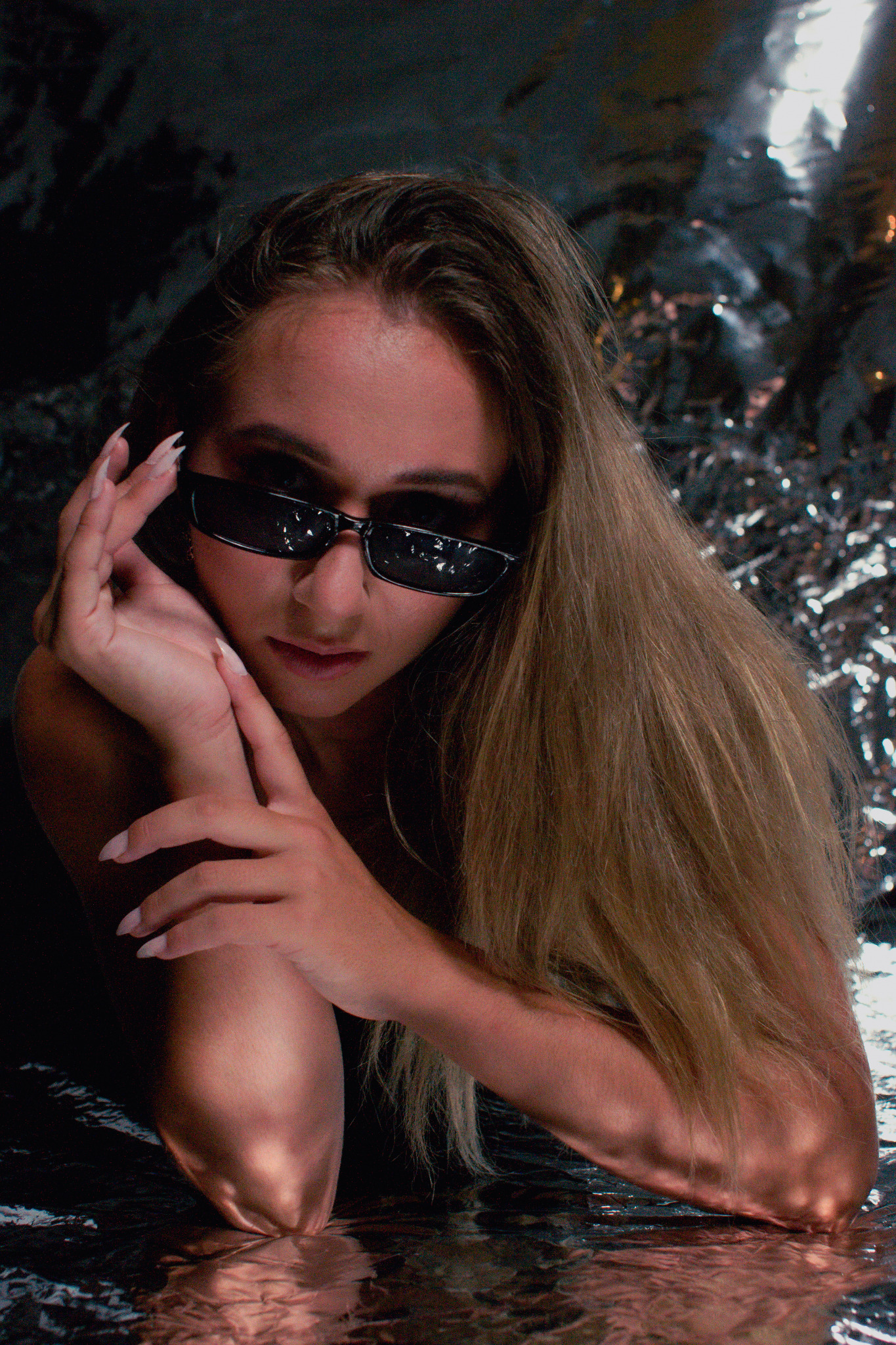 Portret kobiety w okularach przeciwsłonecznych, z dłońmi przy twarzy i długimi paznokciami na tle błyszczącej, srebrnej powierzchni.