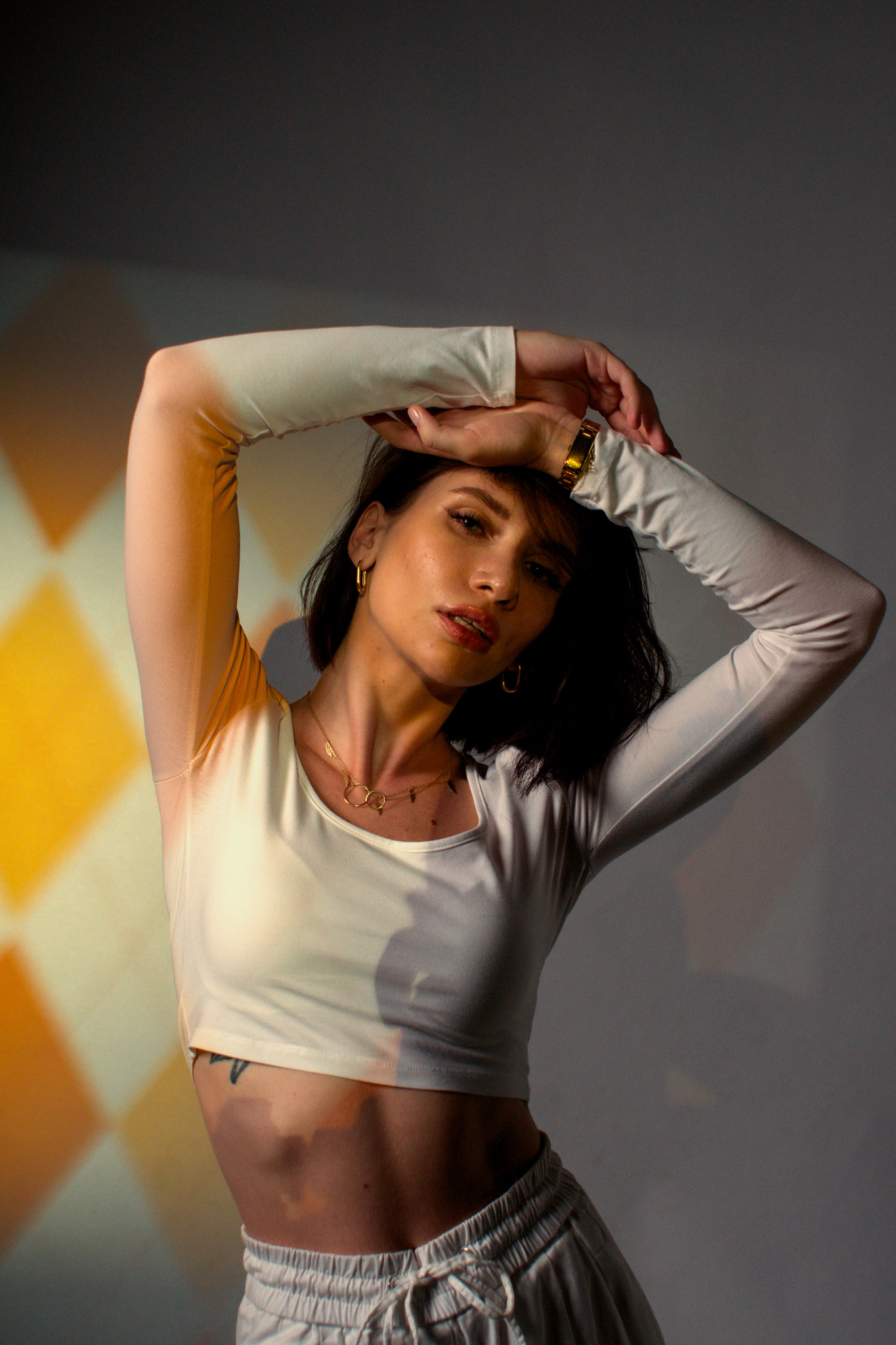 Portret kobiety w białym topie i spodniach dresowych, z rękami uniesionymi nad głową, w studiu z geometrycznym oświetleniem.