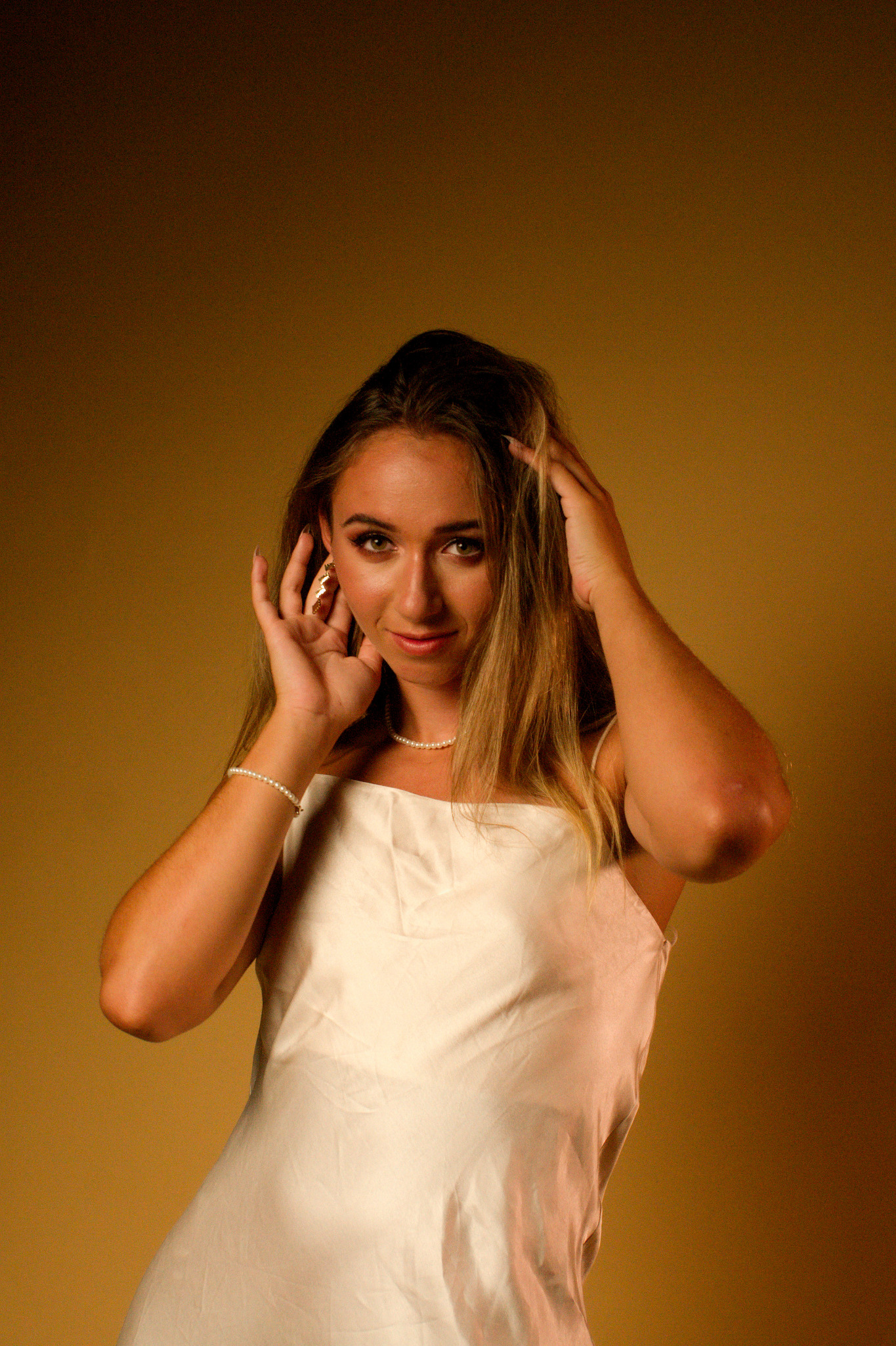 Portret kobiety w białej sukience z perłową biżuterią, pozującej z rękami we włosach na jednolitym, ciemnożółtym tle.