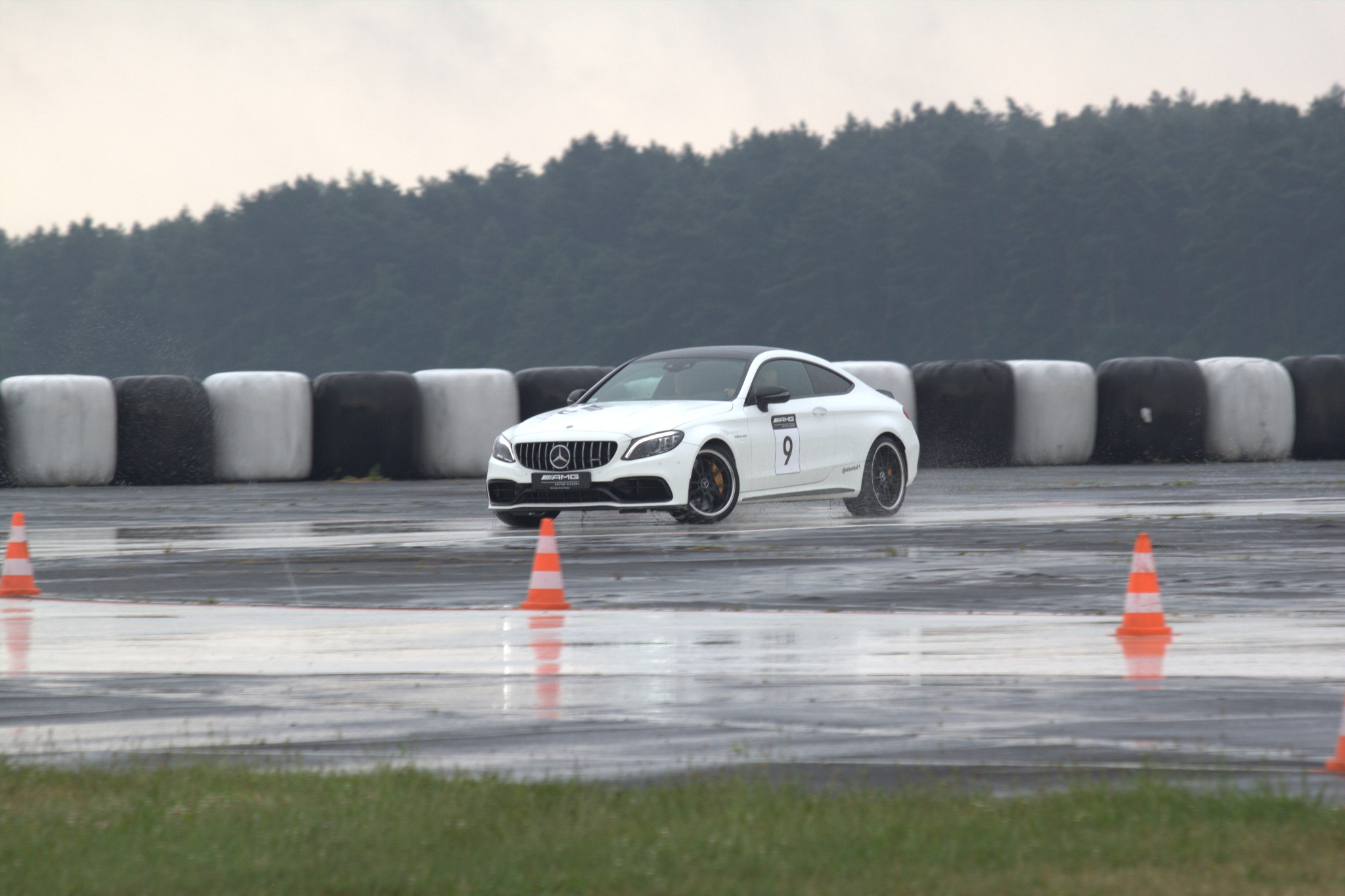 Biały samochód sportowy Mercedes-AMG z numerem 9 na mokrym torze, opony w tle, deszczowa pogoda.