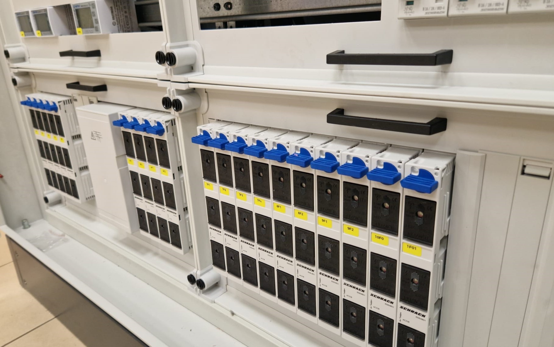 Rozdzielnica elektryczna z modułowymi wyłącznikami firmy Schrack, oznaczonymi żółtymi etykietami, z niebieskimi dźwigniami w pozycji włączonej, zamontowana w białej obudowie z czarnymi uchwytami.
