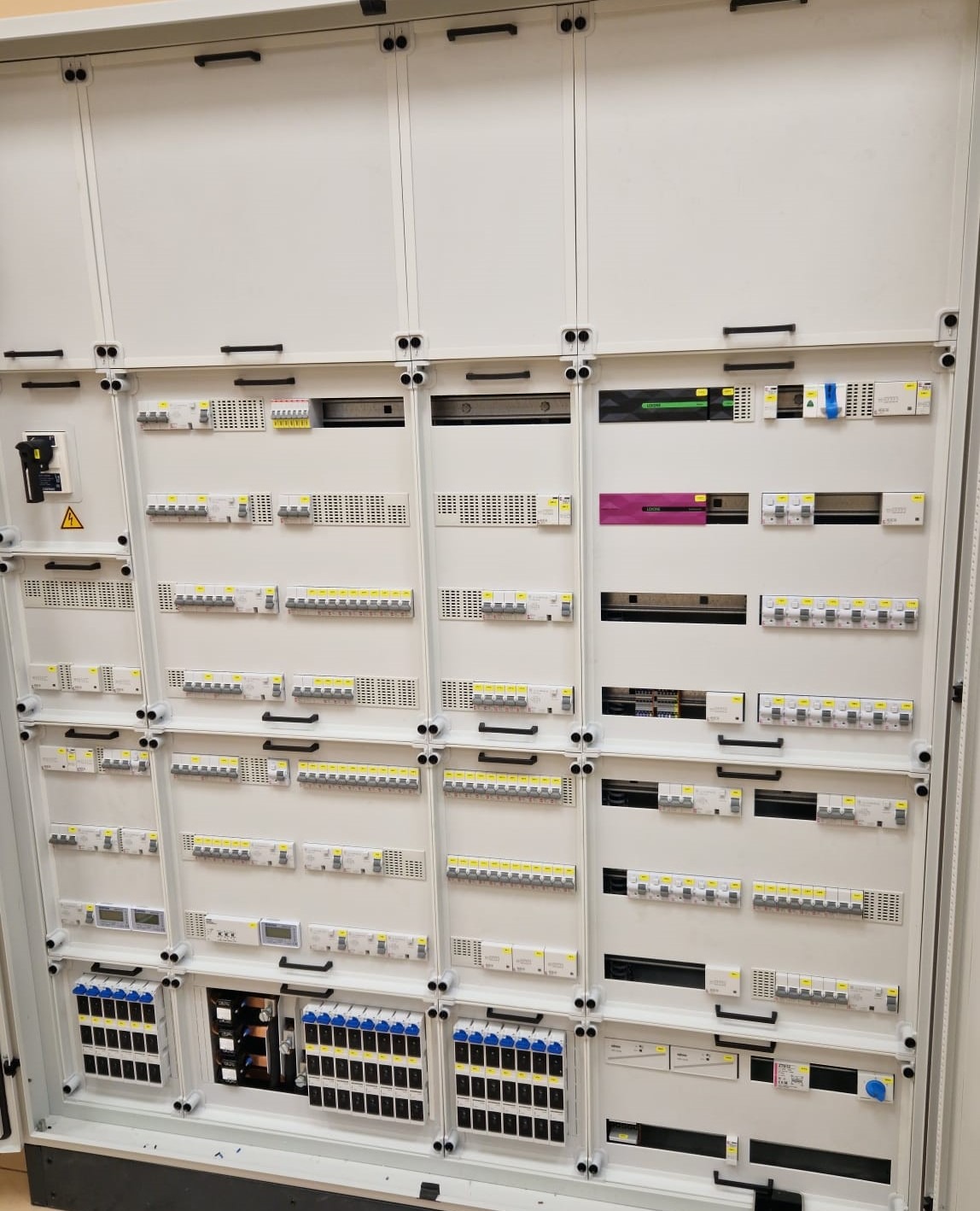 Rozdzielnica elektryczna z licznymi zabezpieczeniami nadprądowymi, wyłącznikami różnicowoprądowymi i modułami pomiarowymi w białej obudowie modułowej.