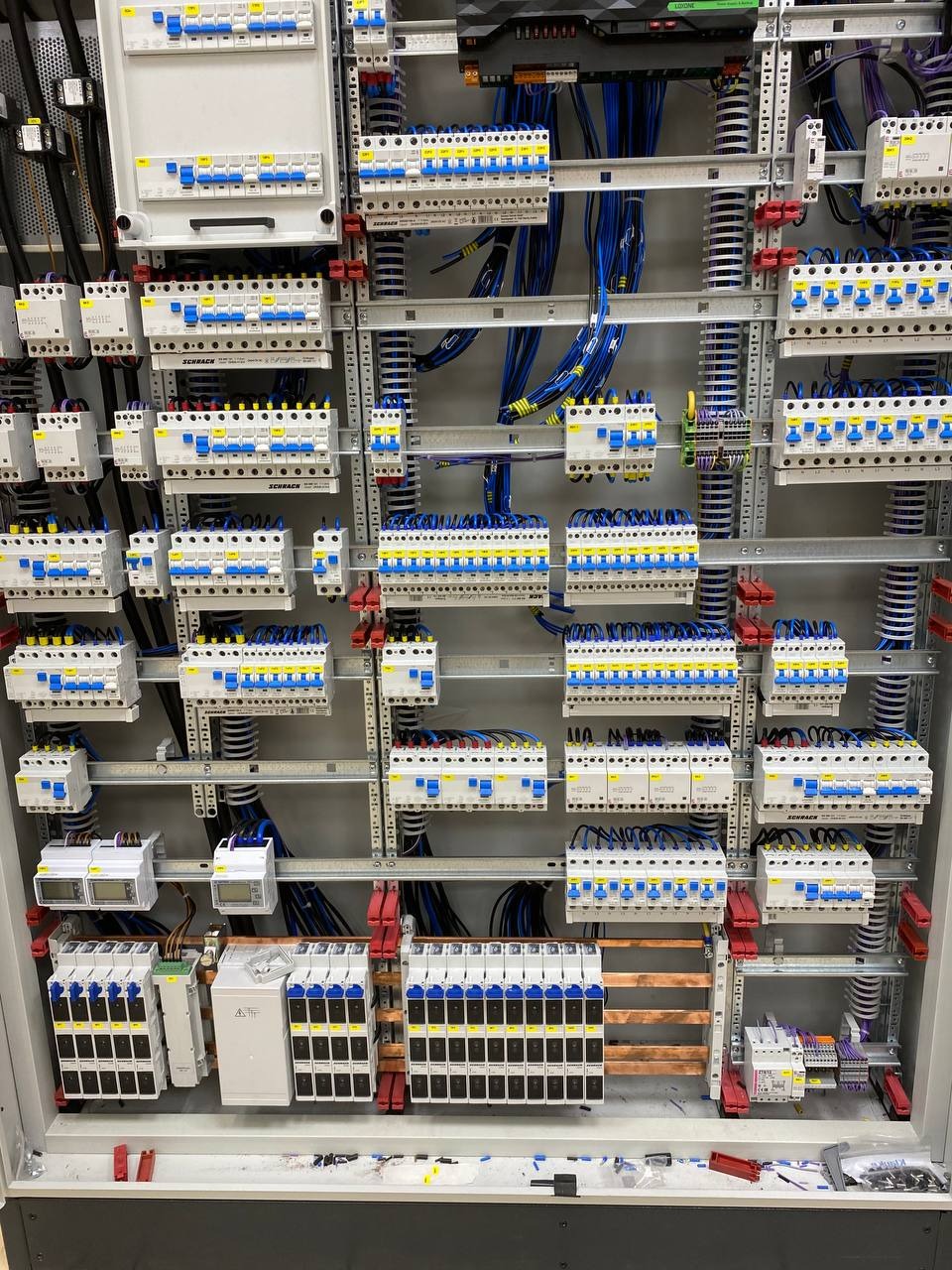 Rozdzielnica elektryczna z licznymi zabezpieczeniami nadprądowymi i przewodami, widoczne szyny montażowe i elementy łączeniowe.