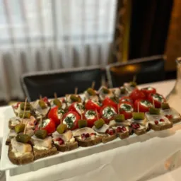 Elegancko ułożone na białym prostokątnym talerzu kanapeczki z pieczywa razowego z śledziem i ogórkiem oraz papryczki nadziewane białym serem i posypane koperkiem, udekorowane ziarnami granatu.