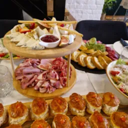 Dwupoziomowa drewniana patera z serami, wędlinami i owocami, obok talerz z roladkami i sałatką, a na pierwszym planie taca z mini tartaletkami z kremowym nadzieniem i czerwoną dekoracją.