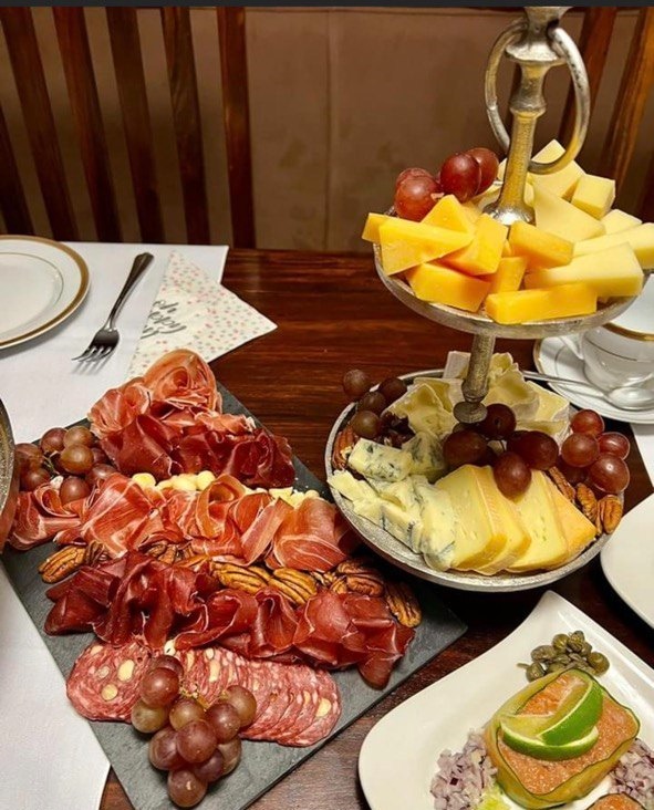 Elegancko ułożone wędliny, sery, orzechy i winogrona na paterach i desce, gotowe do serwowania, obok nakryty stół z talerzami i sztućcami.