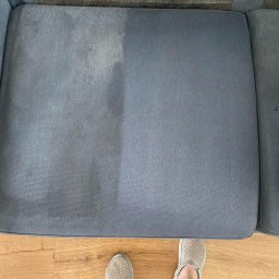 Czysty Sen pranie tapicerki w Lublinie - Widok z góry na fragment szarej kanapy z widoczną różnicą w czystości po praniu; po lewej stronie widoczny ciemniejszy, zabrudzony obszar, po prawej jaśniejszy, wyczyszczony fragment; na dole...