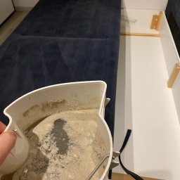 Czysty Sen pranie tapicerki w Lublinie - Pojemnik z brudną wodą po czyszczeniu tapicerki kanapy, widoczna ciemna plama z zanieczyszczeń i resztek detergentu. W tle fragment niebieskiej kanapy z otwieranym schowkiem.
