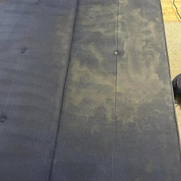 Czysty Sen pranie tapicerki w Lublinie - Granatowa kanapa z widocznymi plamami i różnicą w czystości po praniu, obok leży szczotka do czyszczenia tapicerki.