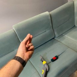 Czysty Sen pranie tapicerki w Lublinie - Dysza odkurzacza piorącego leży na jasnoniebieskiej kanapie z weluru, obok mała szczotka z czerwono-zielonym włosiem, w tle dłoń w geście 'szczypta' i roślina doniczkowa.