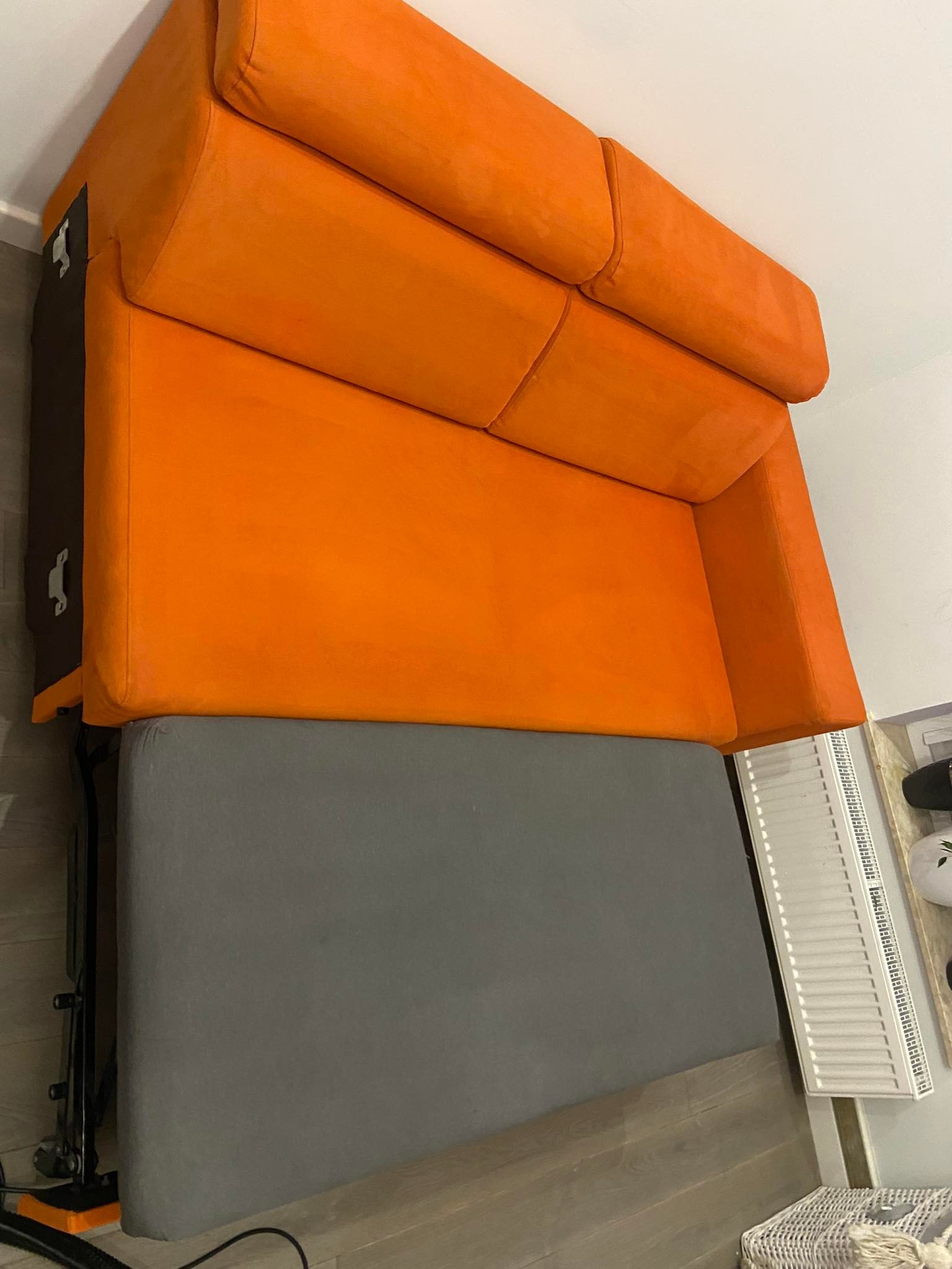 Rozłożona pomarańczowa kanapa z szarą powierzchnią do spania, widoczne elementy mechanizmu rozkładania i fragment podłogi z panelem grzejnikowym w tle.