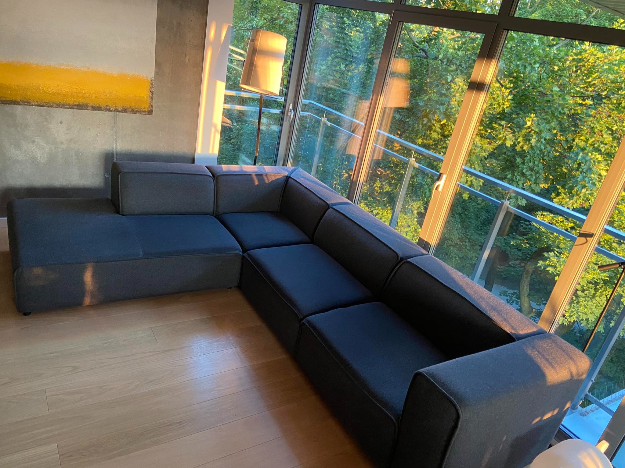 Szara, modułowa sofa w nowoczesnym salonie z dużymi oknami, przez które wpada popołudniowe światło, oświetlając fragment podłogi i mebla, z widokiem na zieleń za oknem.