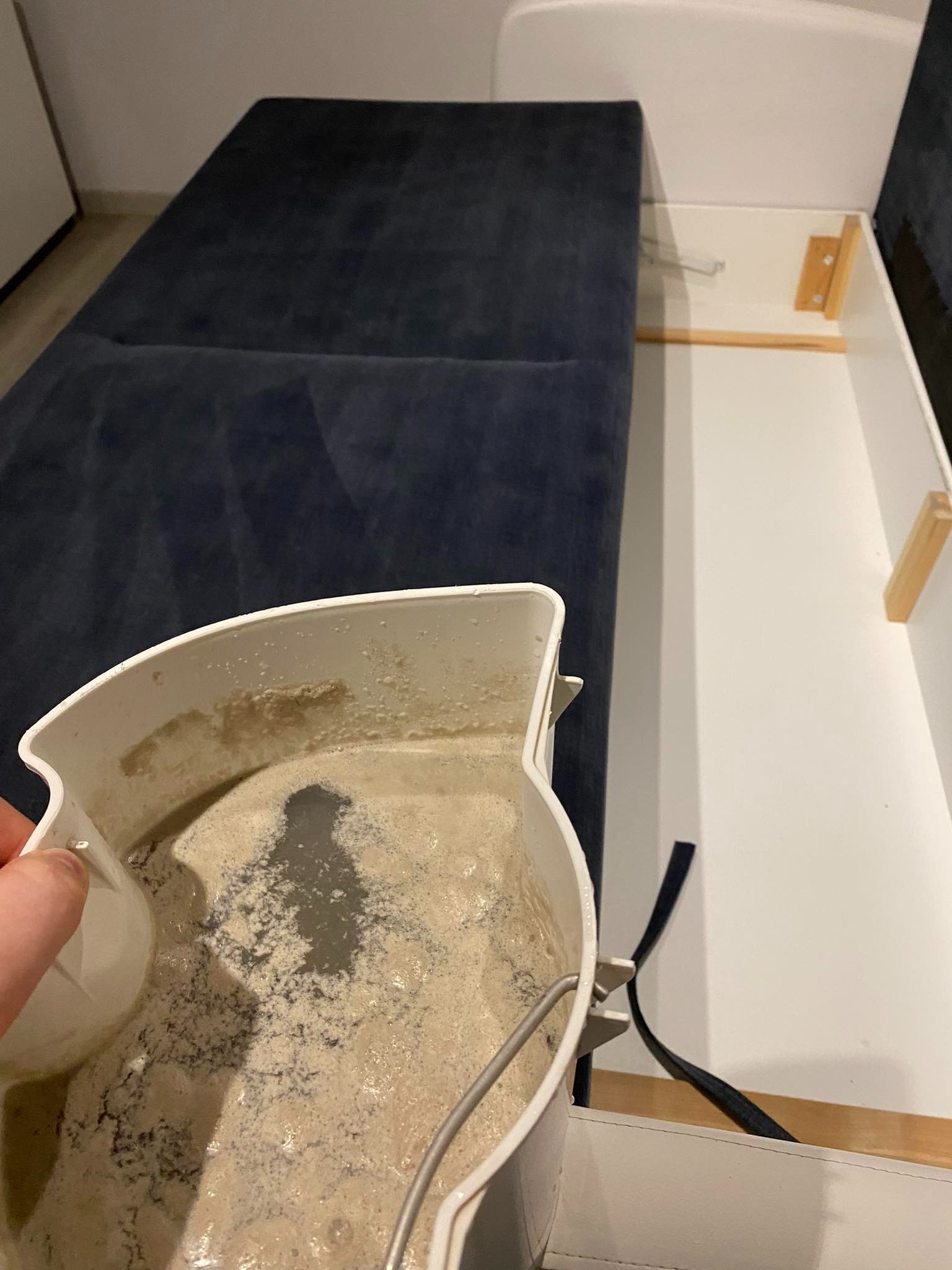 Pojemnik z brudną wodą po czyszczeniu tapicerki kanapy, widoczna ciemna plama z zanieczyszczeń i resztek detergentu. W tle fragment niebieskiej kanapy z otwieranym schowkiem.