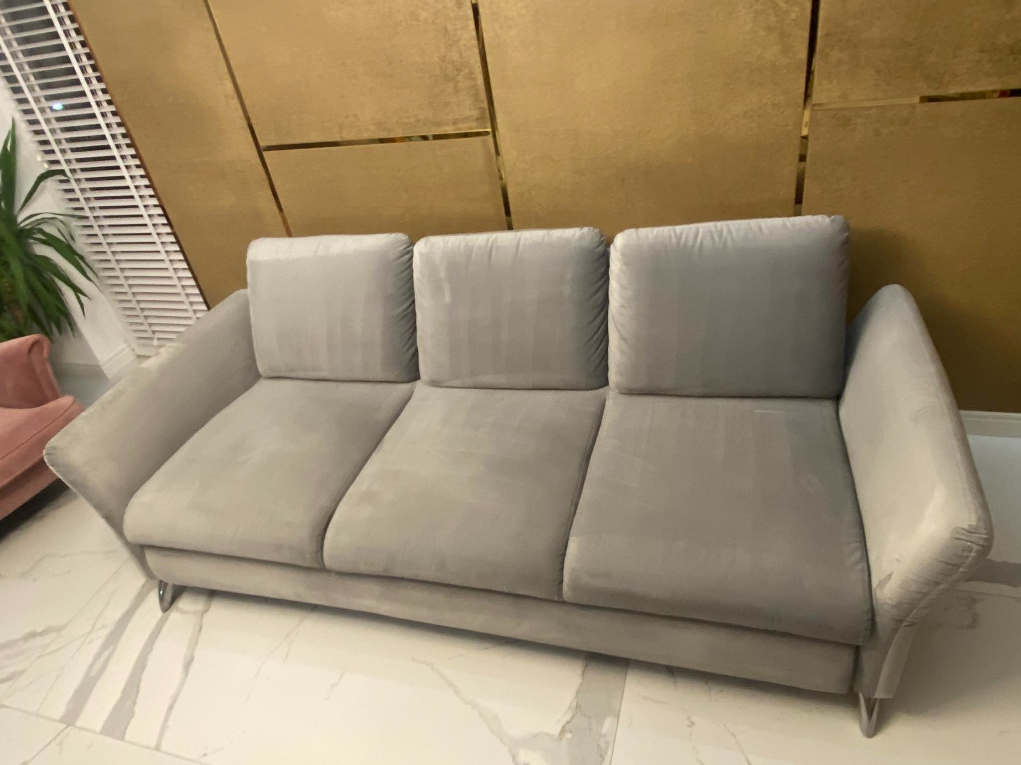 Elegancka, szara, welurowa sofa trzyosobowa z kwadratowymi poduszkami na tle złotej ściany z geometrycznym wzorem i marmurowej podłogi.