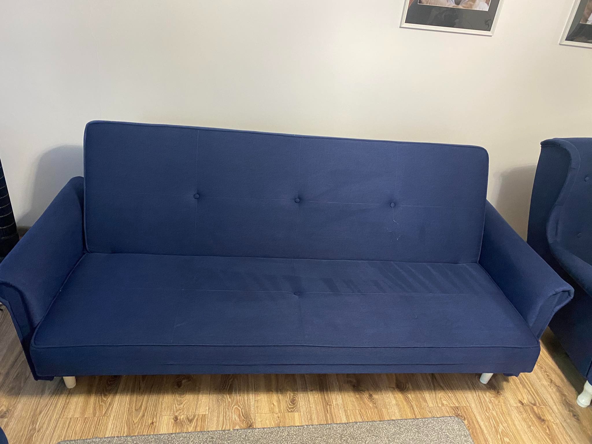 Niebieska kanapa rozkładana z trzema guzikami, ustawiona na drewnianej podłodze, widoczny fragment fotela w tym samym kolorze.