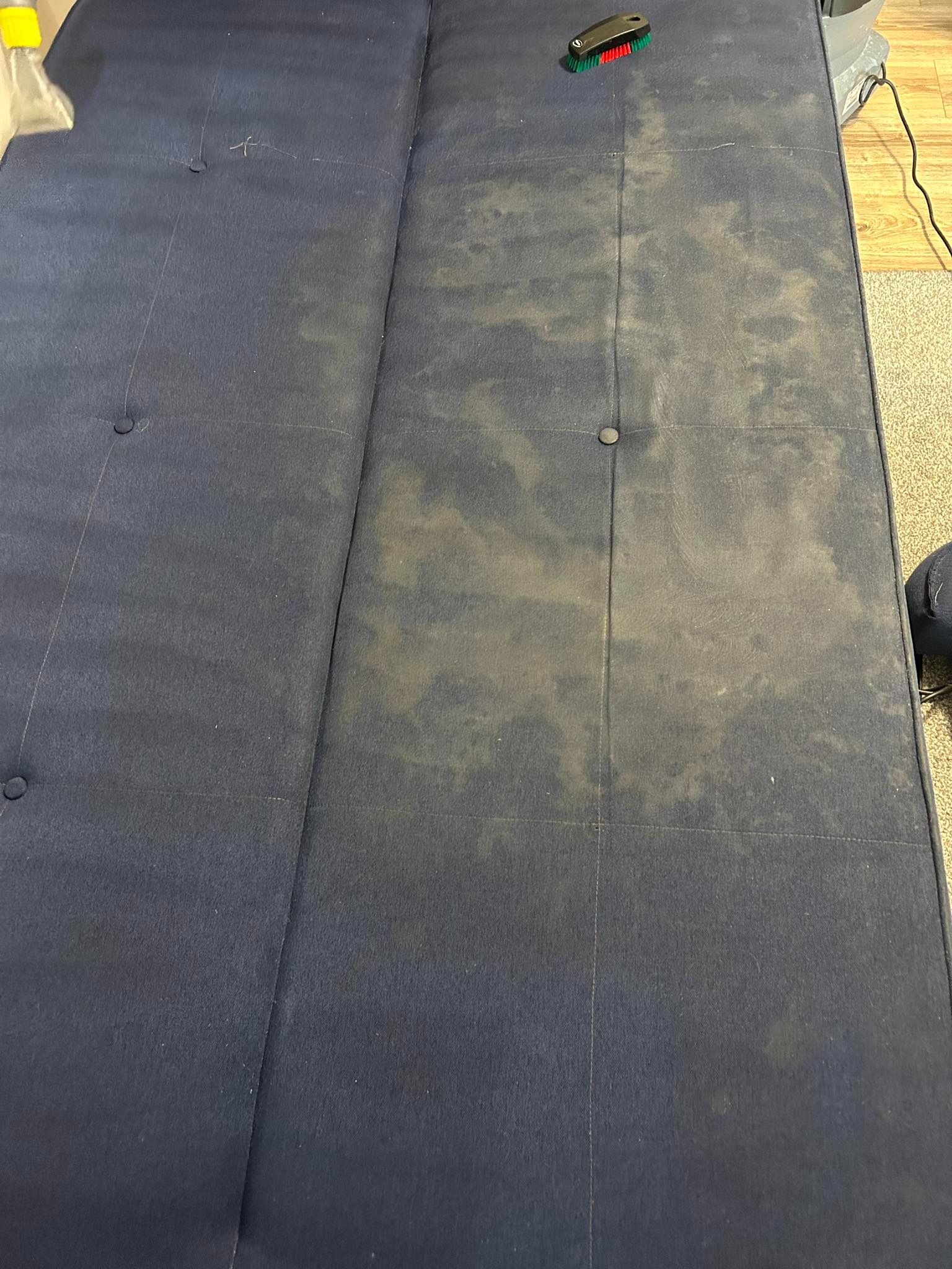 Granatowa kanapa z widocznymi plamami i różnicą w czystości po praniu, obok leży szczotka do czyszczenia tapicerki.