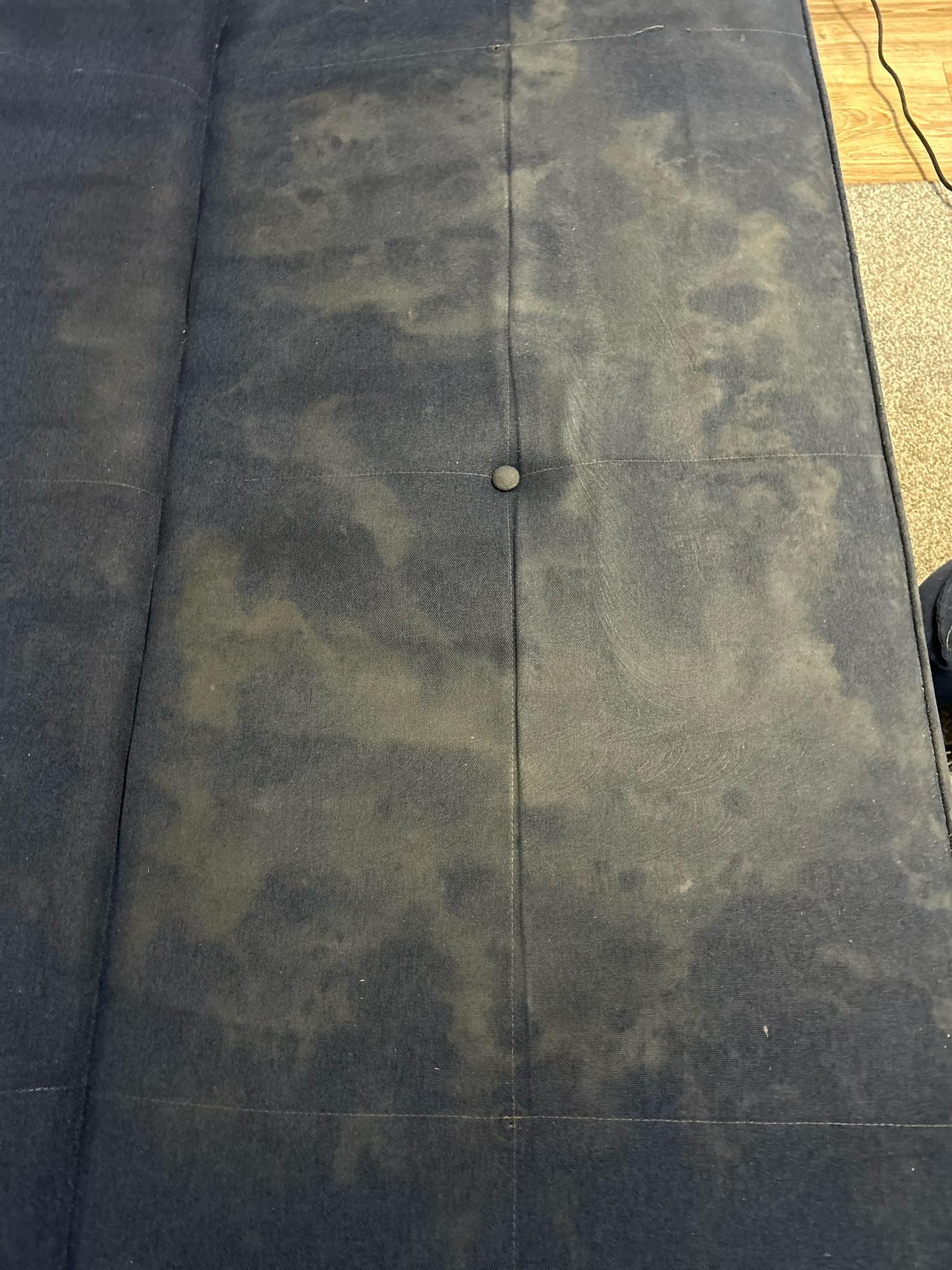 Widok z góry na mocno zabrudzoną, ciemnoniebieską tapicerkę sofy z widocznymi plamami i przebarwieniami, z jednym guzikiem pośrodku i szwami dzielącymi powierzchnię na kwadraty.