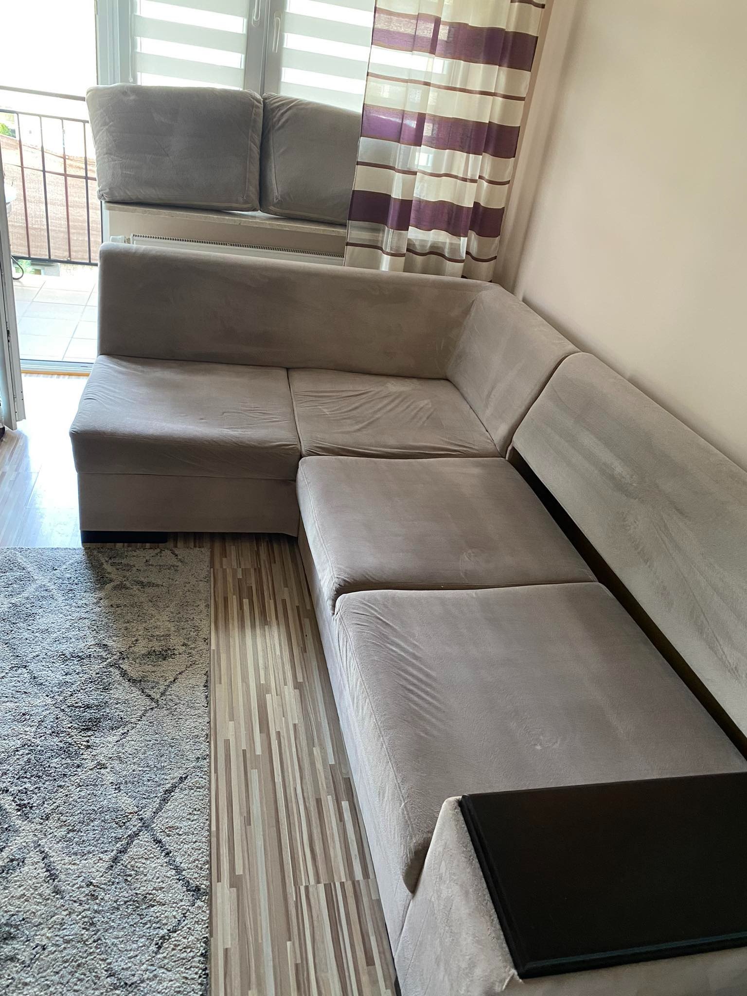 Beżowy narożnik z widocznymi śladami użytkowania, ustawiony w rogu pokoju z dywanem i panelami podłogowymi, oświetlony dziennym światłem z okna z zasłonami w paski.