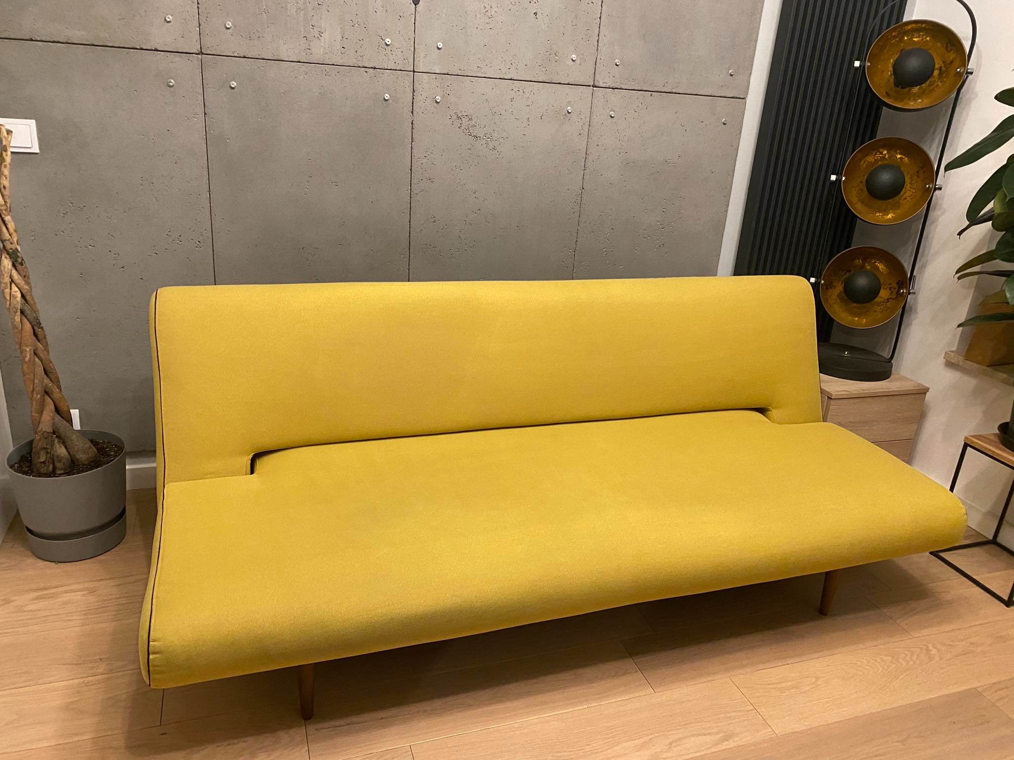 Rozłożona żółta kanapa z tapicerowanymi bokami na drewnianych nóżkach, na tle betonowej ściany z widocznymi mocowaniami, obok doniczka z rośliną i lampa podłogowa z trzema okrągłymi kloszami.