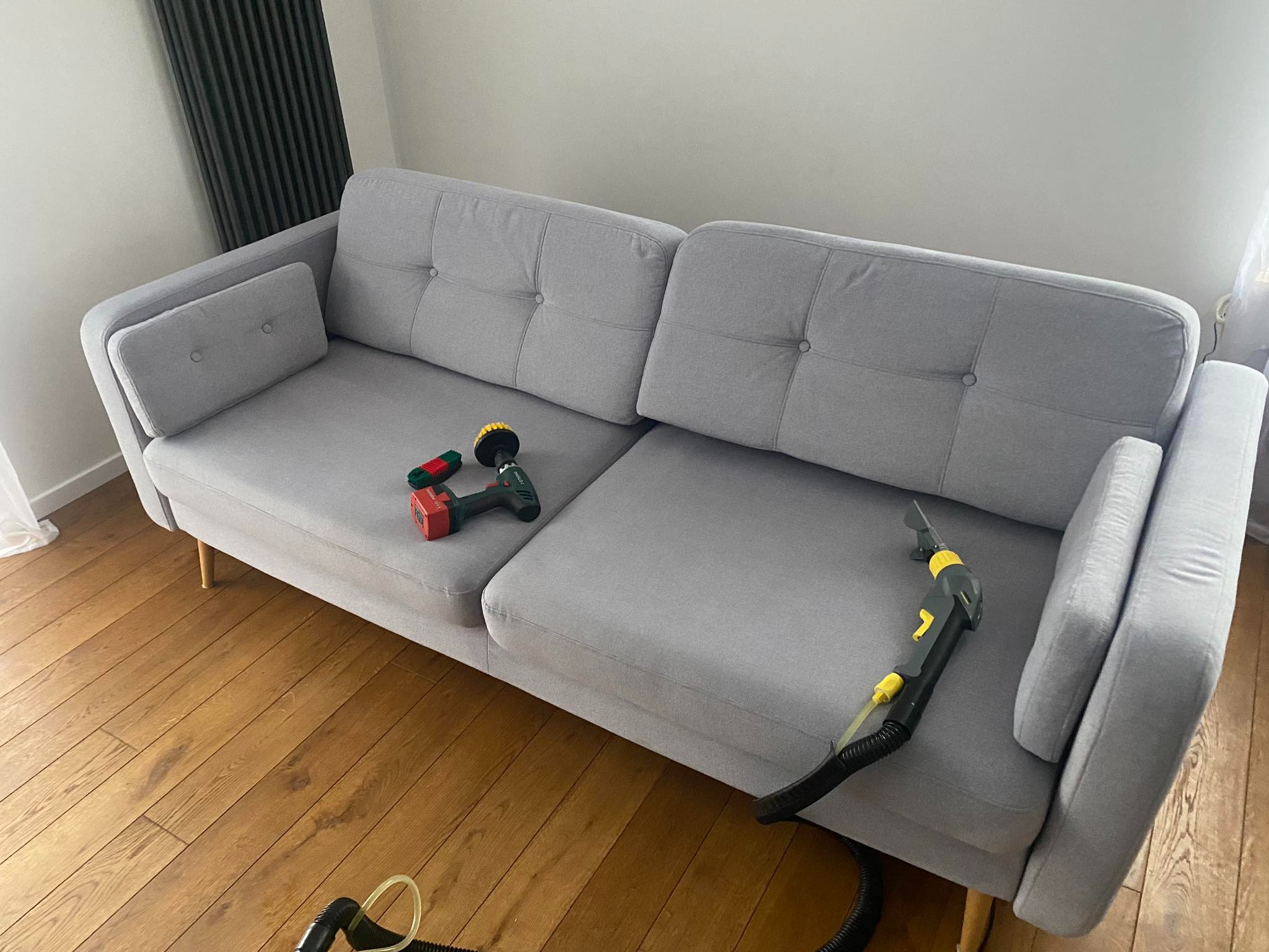 Szara sofa z guzikami na siedzisku, czyszczona parownicą z żółtymi elementami, na podłodze leży wiertarka z szczotką, widoczny grzejnik.
