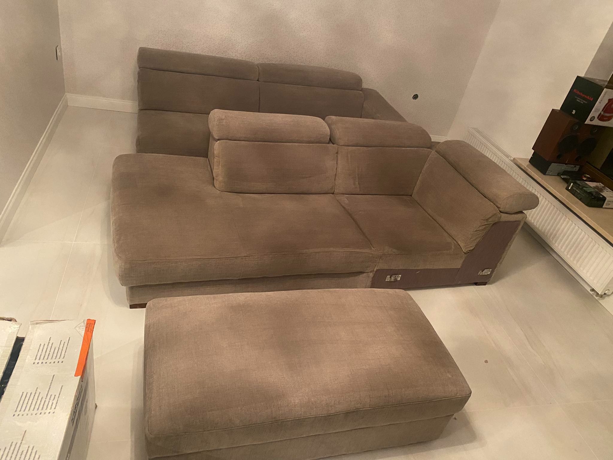 Beżowy narożnik z pufą na białych płytkach, obok kartonowe pudła, w tle grzejnik i pudełka na półce.