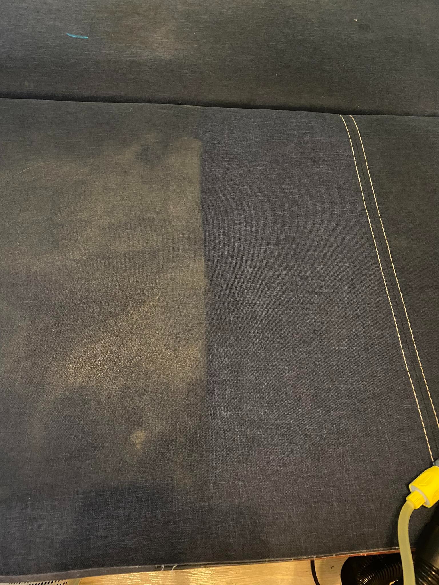 Fragment ciemnej tapicerki kanapy, widoczny efekt czyszczenia: lewa strona jaśniejsza, prawa strona ciemniejsza, z żółtą końcówką węża do prania w prawym dolnym rogu.