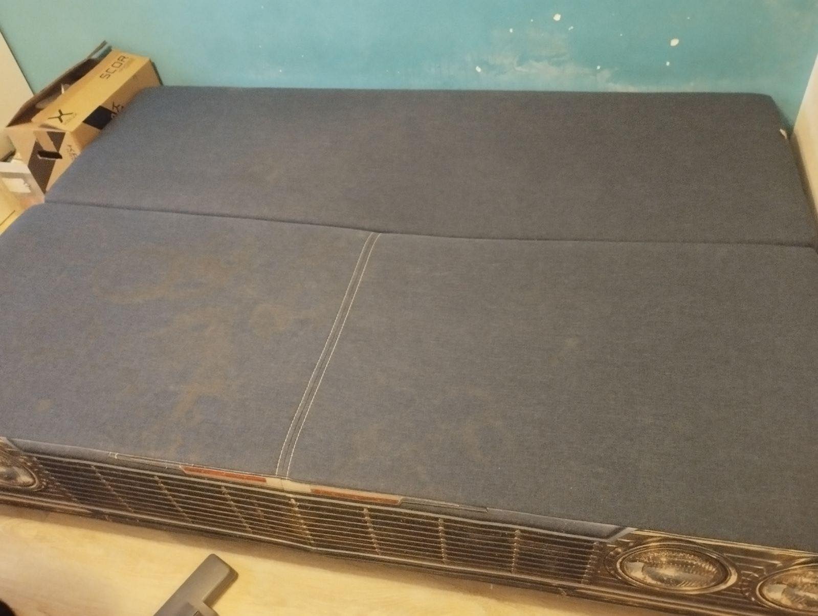 Szara, dwuczęściowa kanapa z wyraźnymi plamami na tapicerce, z dekoracyjnym panelem imitującym przód samochodu, widoczny fragment odkurzacza na podłodze.