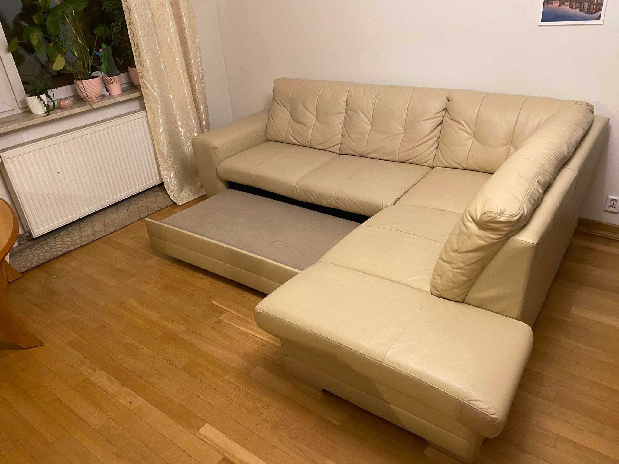 Rozłożona beżowa kanapa narożna z funkcją spania na parkiecie, widok z boku, z roślinami w doniczkach na parapecie w tle.