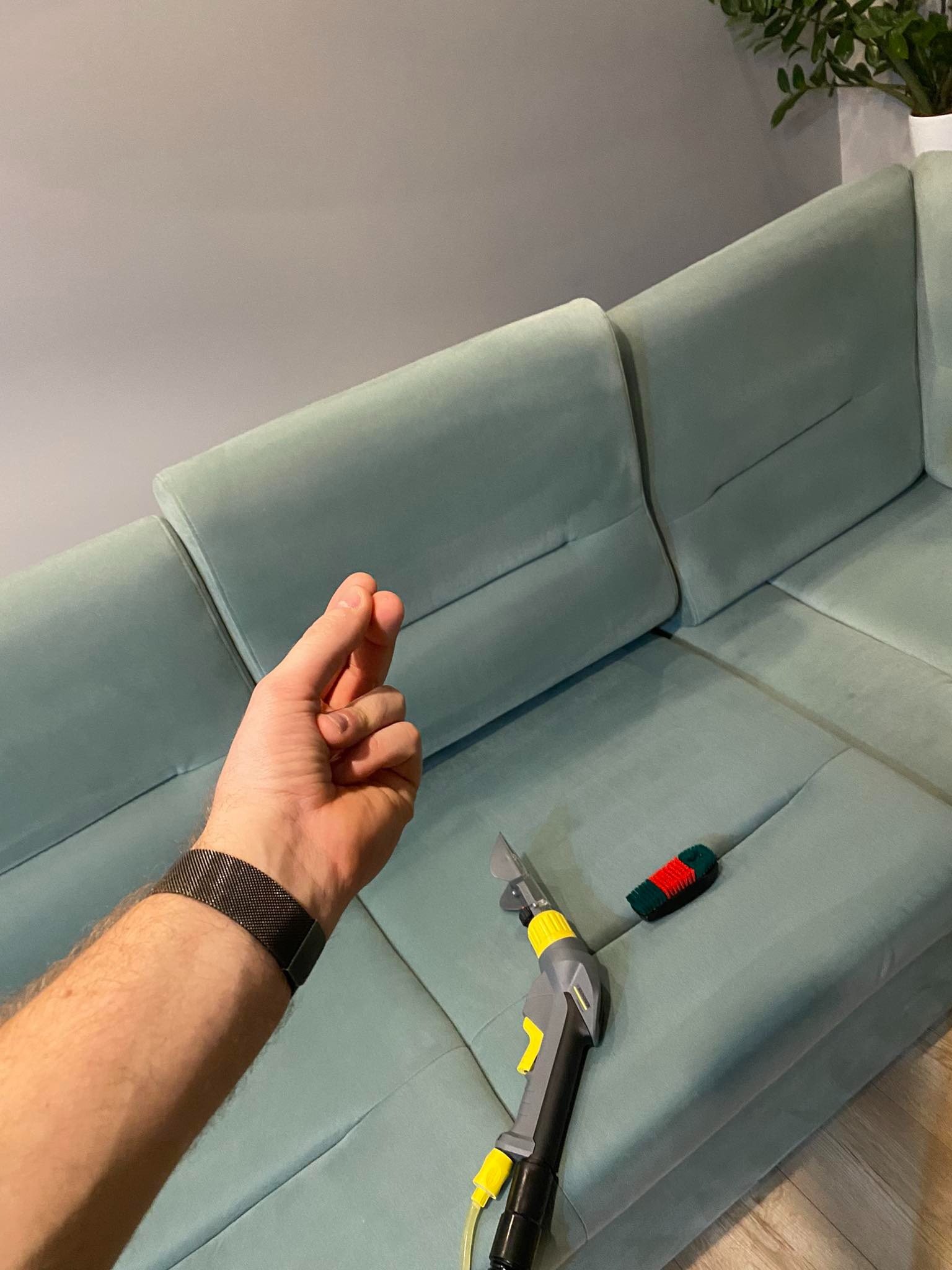 Dysza odkurzacza piorącego leży na jasnoniebieskiej kanapie z weluru, obok mała szczotka z czerwono-zielonym włosiem, w tle dłoń w geście 'szczypta' i roślina doniczkowa.