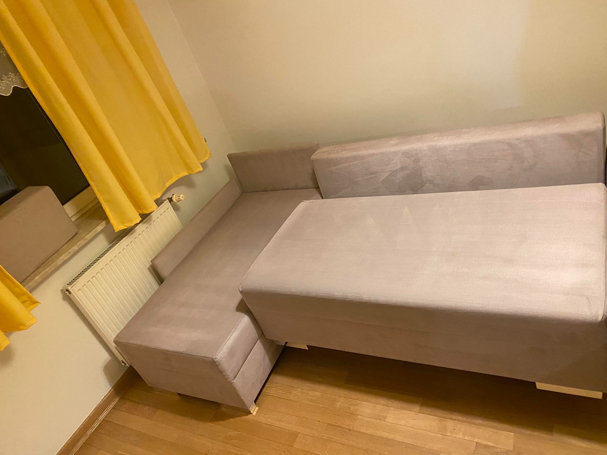 Beżowa, modułowa kanapa z aksamitną tapicerką, ustawiona na drewnianej podłodze, z widocznym fragmentem żółtej zasłony i białego grzejnika.