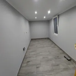 Pomalowane na szaro wnętrze pokoju z szarą podłogą z płytek imitujących drewno, oświetlone sześcioma punktowymi lampami sufitowymi, z oknem po prawej stronie i wystającymi przewodami elektrycznymi...