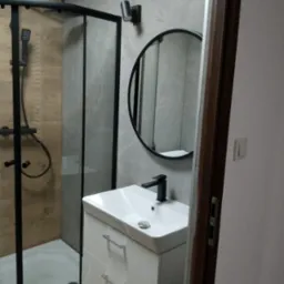 Nowoczesna łazienka z kabiną prysznicową z czarną ramą, okrągłym lustrem w czarnej ramie, białą szafką pod umywalką i szarą płytką imitującą kamień na ścianach