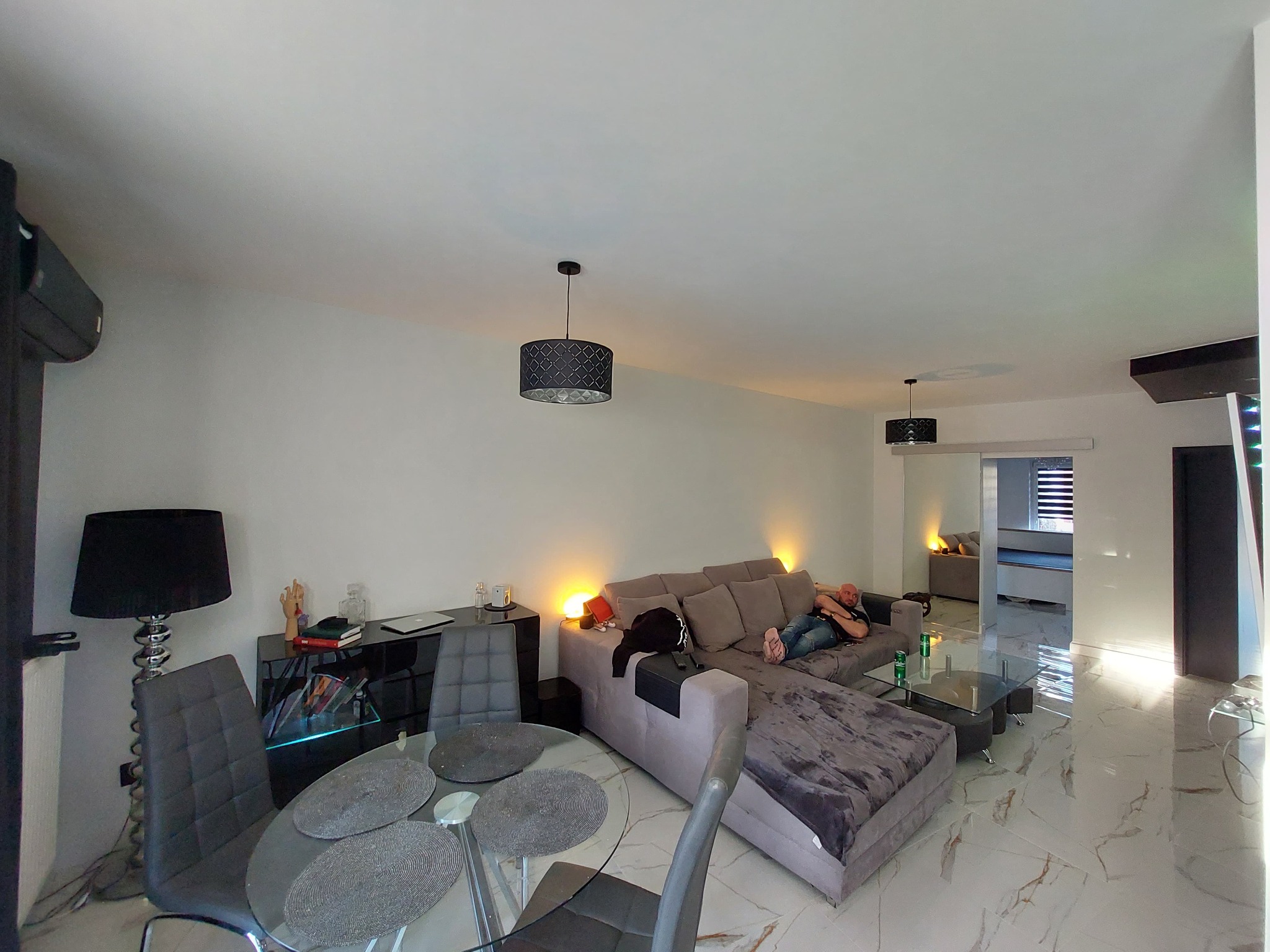 Jasne, przestronne wnętrze salonu z marmurową podłogą, szarym narożnikiem, szklanym stołem i lampami wiszącymi, oświetlone ciepłym światłem.